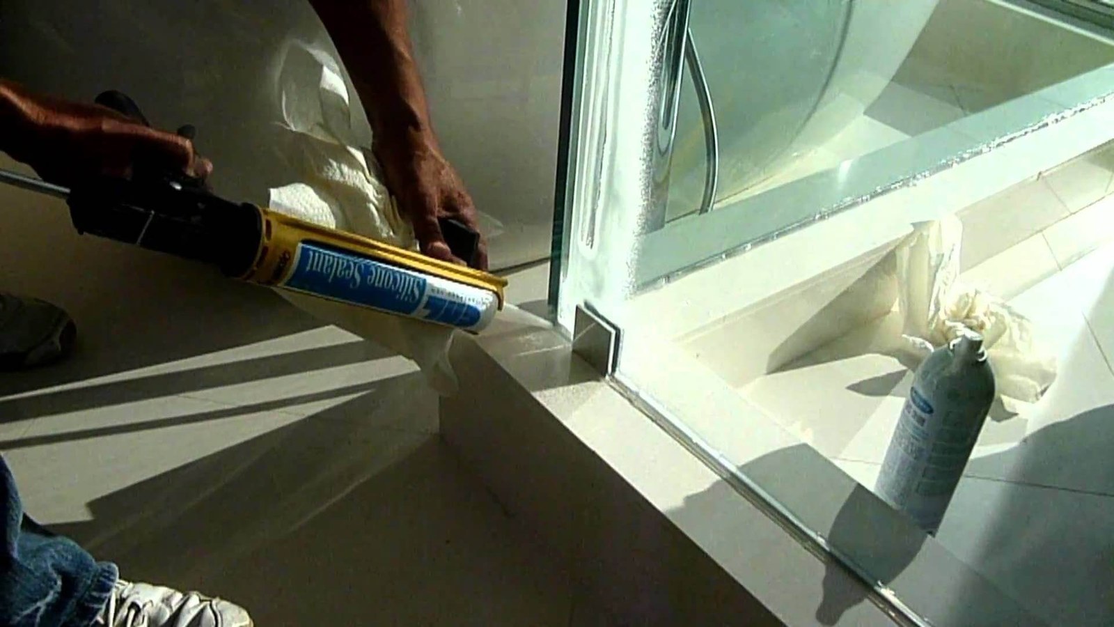 A glazier applying silicone sealant along the base of a glass shower panel on a tiled floor
